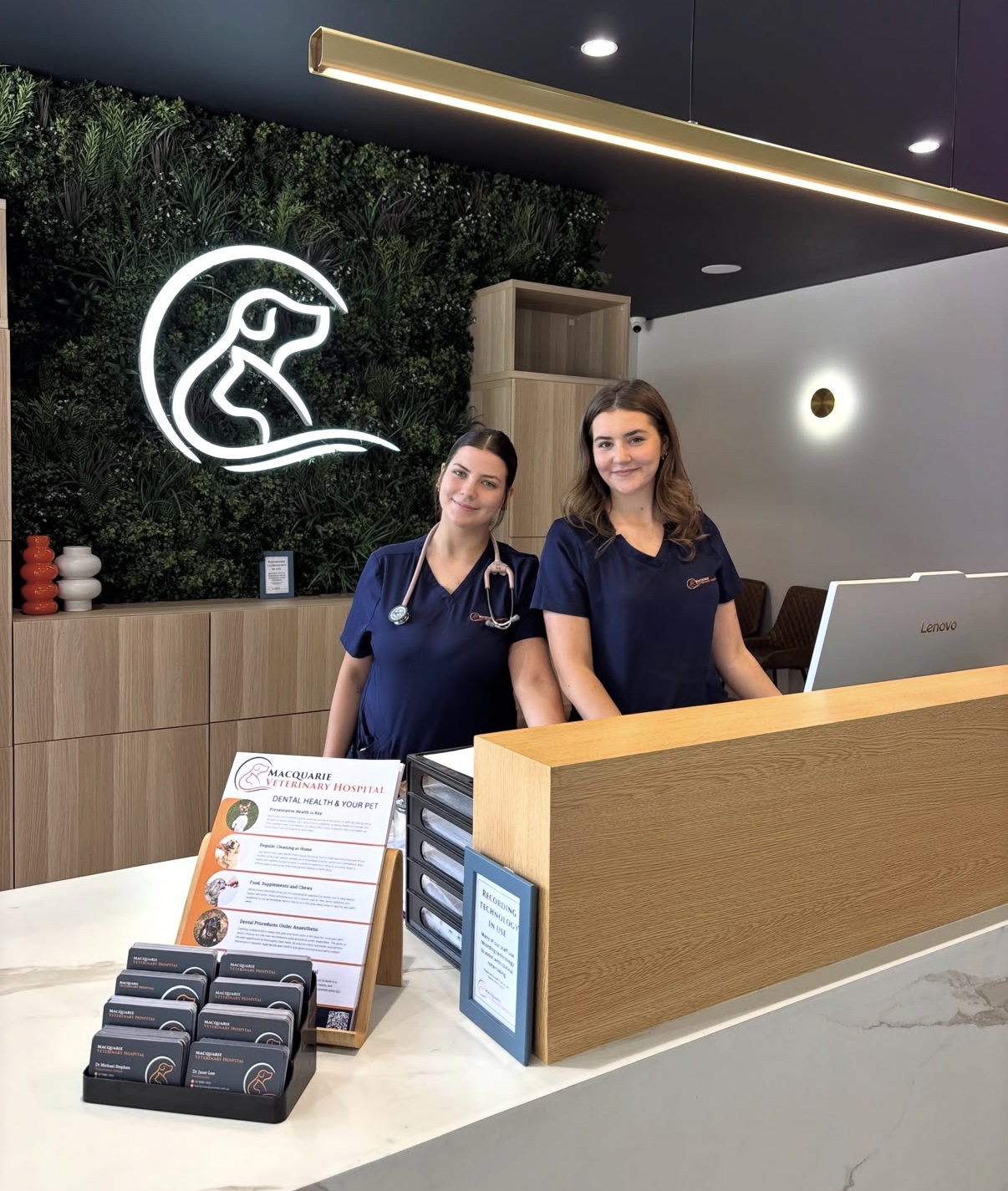 Two members of the Macquarie Veterinary Hospital team behind the reception counter at the new hospital, in front of the illuminated logo on a green living wall